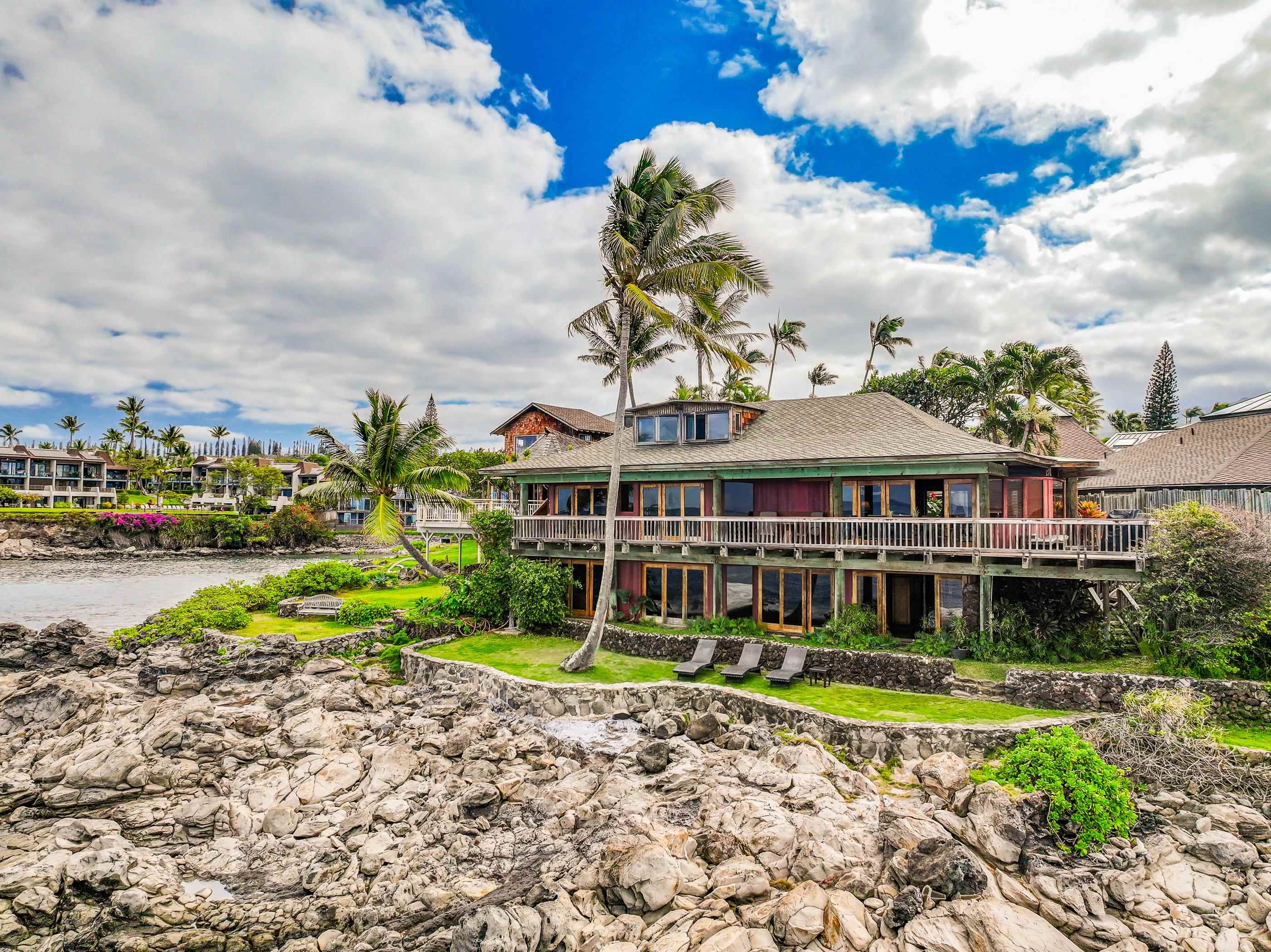 The oceanfront home from the lava-rock shoreline, lounge chairs on the lawn beneath a tall coconut palm and the deck wrapping the upper floor
